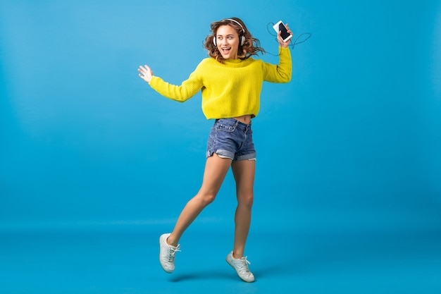 Atractiva mujer feliz sonriente saltando bailando escuchando música en auriculares en traje hipster aislado sobre fondo azul de estudio, vistiendo pantalones cortos y suéter amarillo