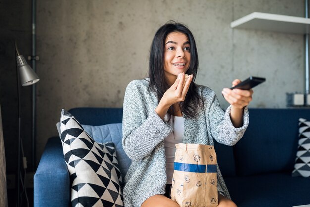 Atractiva mujer feliz sonriente sentada en el sofá en casa viendo películas de televisión en línea con control remoto comiendo palomitas de maíz con expresión de la cara de sorpresa emocional