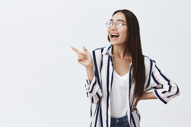 Atractiva mujer femenina con elegante blusa y gafas, riendo a carcajadas, señalando y mirando a la esquina superior izquierda