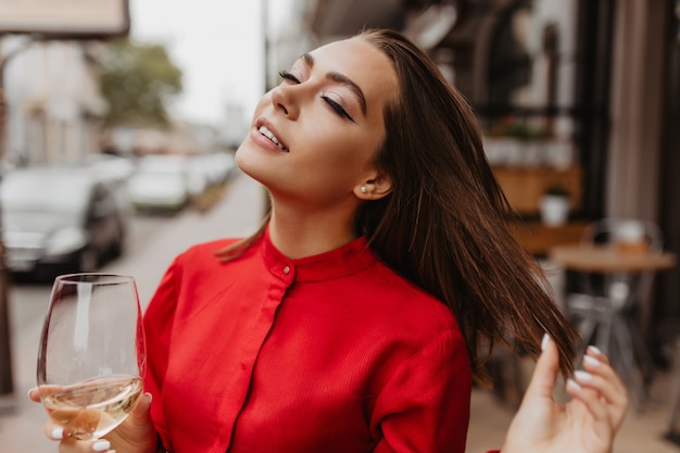 Atractiva mujer francesa en elegante blusa roja disfrutando del sabor del champán. Retrato al aire libre de morena con pelo corto y liso