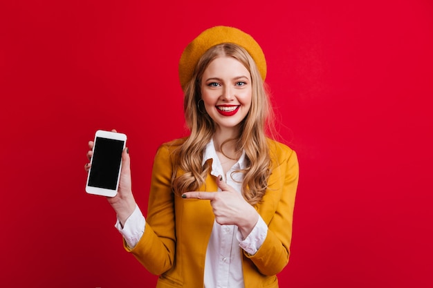 Atractiva mujer francesa sosteniendo smartphone con pantalla en blanco. Vista frontal de la niña de boina amarilla apuntando con el dedo al dispositivo digital.