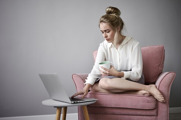 Atractiva mujer independiente en blusa blanca trabajando de forma remota en una computadora portátil, sentada descalza en un sillón, bebiendo té o café caliente, manteniendo una mano en el teclado, teniendo una mirada enfocada
