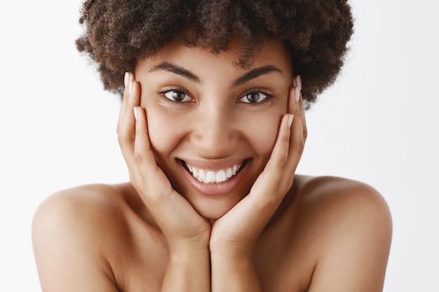 Atractiva mujer joven afroamericana femenina y natural con cabello rizado y piel pura y limpia, tocando la cara y sonriendo ampliamente con entusiasmo y felicidad, posando desnuda sobre una pared gris