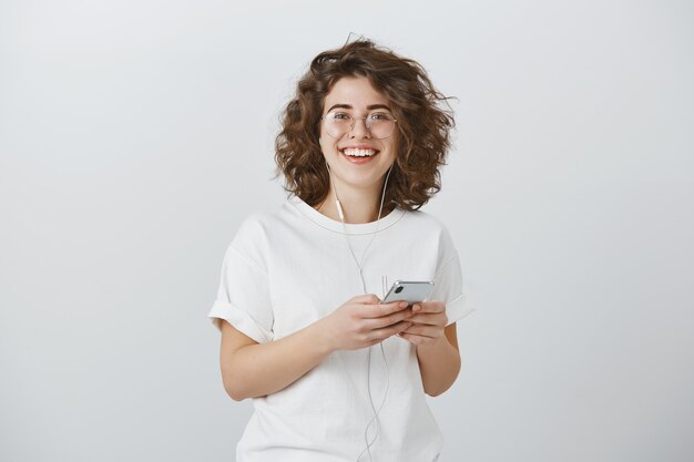 Atractiva mujer joven alegre con gafas, sonriendo mientras sostiene el teléfono inteligente y escucha música en auriculares
