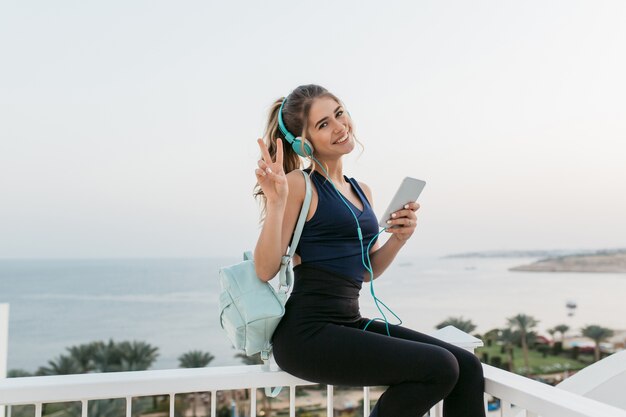 Atractiva mujer joven alegre en ropa deportiva sonriendo, charlando por teléfono, relajándose en el paseo marítimo. Amanecer por la mañana, modelo de moda, entrenamiento, música en auriculares, estado de ánimo alegre