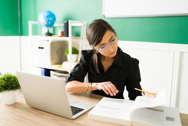 Atractiva mujer joven con anteojos estudiando mientras planifica su clase con libros de texto y su computadora portátil en casa