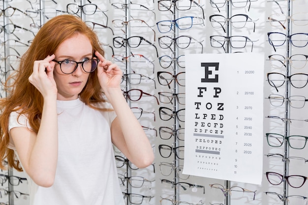 Atractiva mujer joven con anteojos de pie ordenado gráfico de snellen en optica