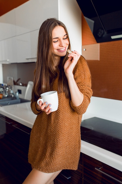 Atractiva mujer joven y bonita con una taza de té blanco mirando por la cámara en la cocina moderna en la soleada mañana de otoño.