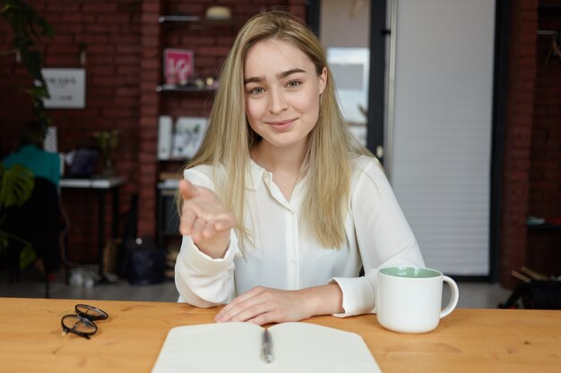 Atractiva mujer joven con cabello rubio suelto sentado en la mesa de café con taza, vasos y cuaderno abierto sobre la mesa, gesticulando con la mano, apuntando al asiento vacante y sonriendo amigablemente