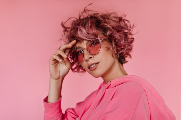 Atractiva mujer joven con capucha rosa y gafas de sol mira a la cámara y toca el cabello La chica bonita sonríe en un fondo aislado