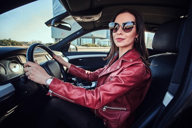 Atractiva mujer joven con chaqueta roja y gafas de sol conduciendo su auto.