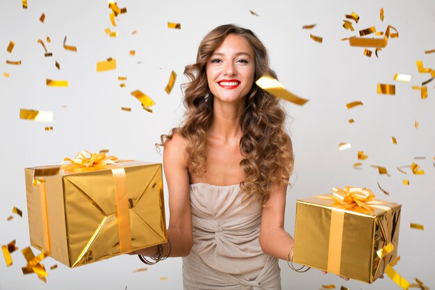 Atractiva mujer joven y elegante celebrando el año nuevo, con regalos en caja, confeti dorado volando, sonriendo feliz, vestido de fiesta, maquillaje de lujo y peinado