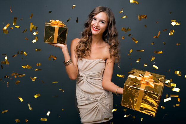 Atractiva mujer joven y elegante celebrando el año nuevo, con regalos en caja, confeti dorado volando, sonriendo feliz, vestido de fiesta, maquillaje de lujo y peinado