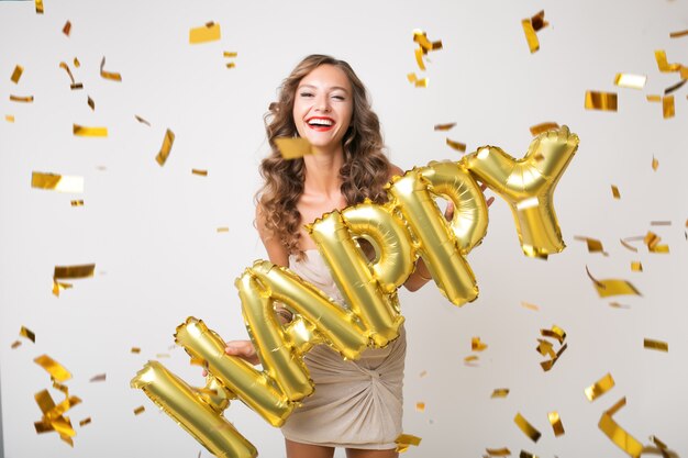 Atractiva mujer joven y elegante celebrando el año nuevo, sosteniendo globos de aire letras felices, confeti dorado volando, sonriendo feliz, aislado, vestido de fiesta