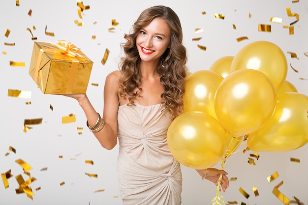 Atractiva mujer joven y elegante celebrando el año nuevo, sosteniendo globos de aire y presenta sorpresa, confeti dorado volando, sonriendo feliz, aislado, vestido de fiesta