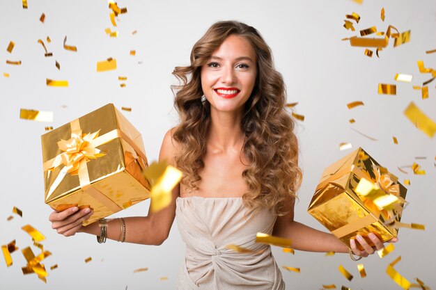 Atractiva mujer joven y elegante celebrando el año nuevo, sosteniendo regalos en caja, confeti dorado volando, sonriendo feliz, vestido de fiesta