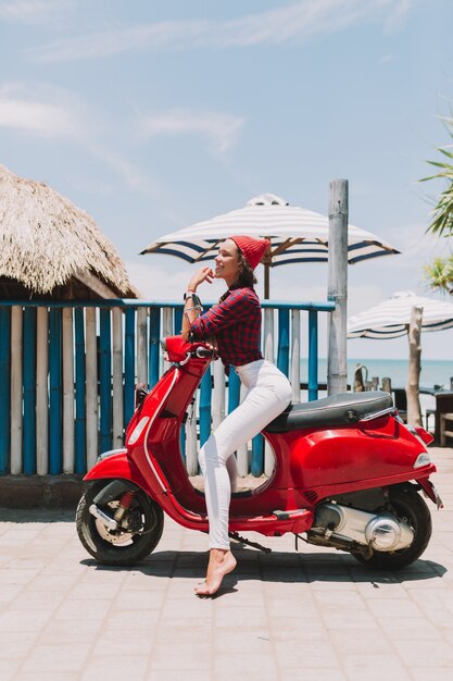 Atractiva mujer joven elegante vestida con pantalones blancos y camisa con gafas de sol posando mientras está sentado en la moto roja junto al océano