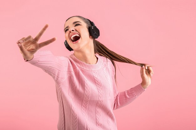 Atractiva mujer joven escuchando música en auriculares inalámbricos con suéter rosa sonriendo feliz estado de ánimo positivo posando sobre fondo rosa