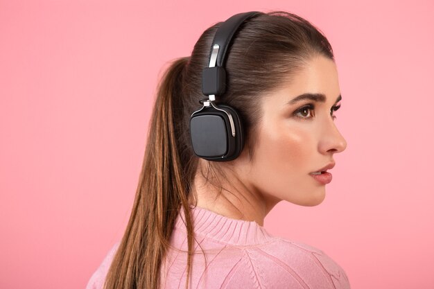 Atractiva mujer joven escuchando música en auriculares inalámbricos con suéter rosa sonriendo feliz estado de ánimo positivo posando sobre fondo rosa