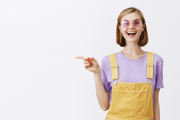 Atractiva mujer joven con estilo en gafas de sol sonriendo, recomendar promoción, señalar con el dedo a la izquierda en copyspace