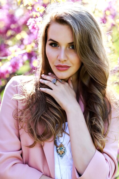 Atractiva mujer joven con estilo en vestido blanco claro, abrigo rosa, con cabello largo caminando en el jardín con sakura floreciente