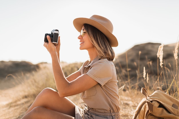 Atractiva mujer joven con estilo en vestido caqui en el desierto, viajando en África en un safari, con sombrero y mochila, tomando fotos con una cámara vintage, explorando la naturaleza, clima soleado