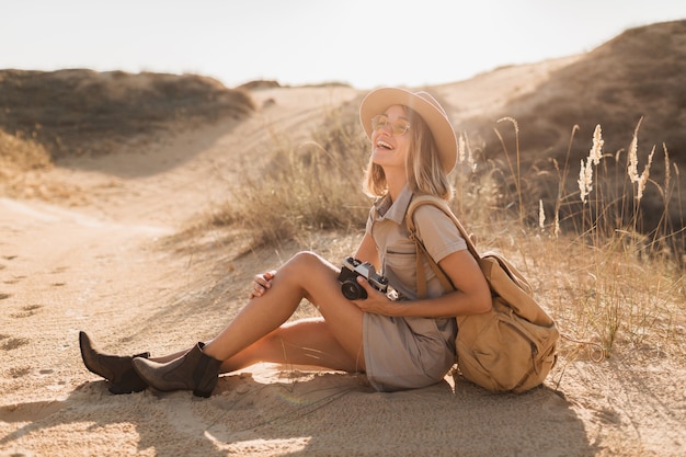 Atractiva mujer joven con estilo en vestido caqui en el desierto, viajando en África en un safari, con sombrero y mochila, tomando fotos con una cámara vintage