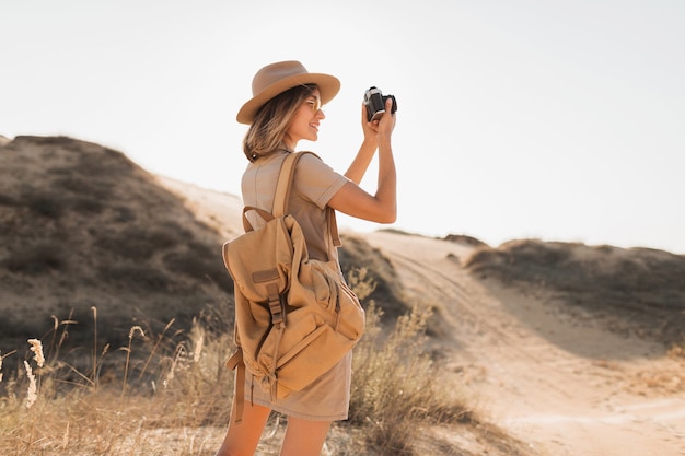 Atractiva mujer joven con estilo en vestido caqui en el desierto, viajando en África en un safari, con sombrero y mochila, tomando fotos con una cámara vintage