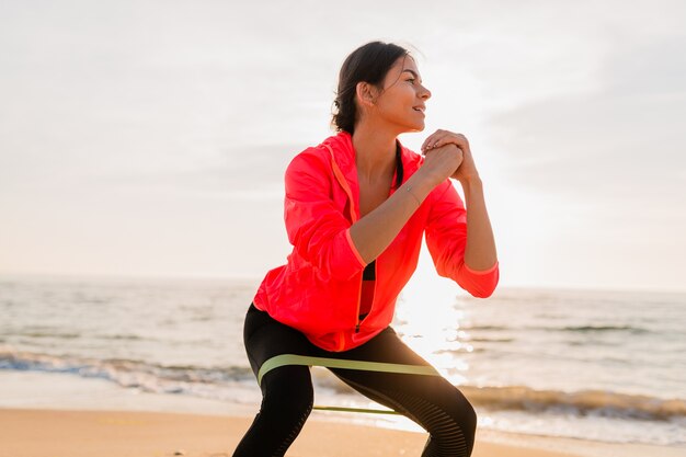 Atractiva mujer joven haciendo ejercicios deportivos en el amanecer de la mañana en la playa del mar, estilo de vida saludable, escuchando música en auriculares, vistiendo una chaqueta cortavientos rosa, haciendo estiramientos en goma elástica