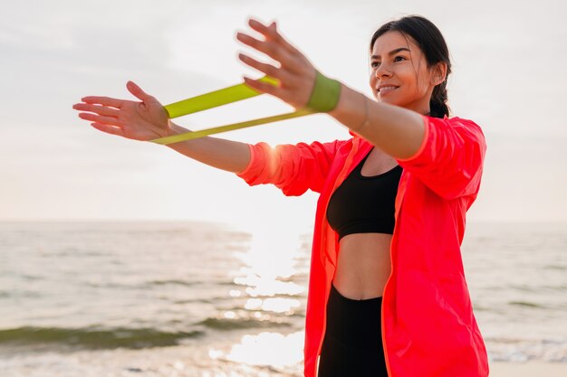 Atractiva mujer joven haciendo ejercicios deportivos en el amanecer de la mañana en la playa del mar, estilo de vida saludable, escuchando música en auriculares, vistiendo una chaqueta cortavientos rosa, haciendo estiramientos en goma elástica