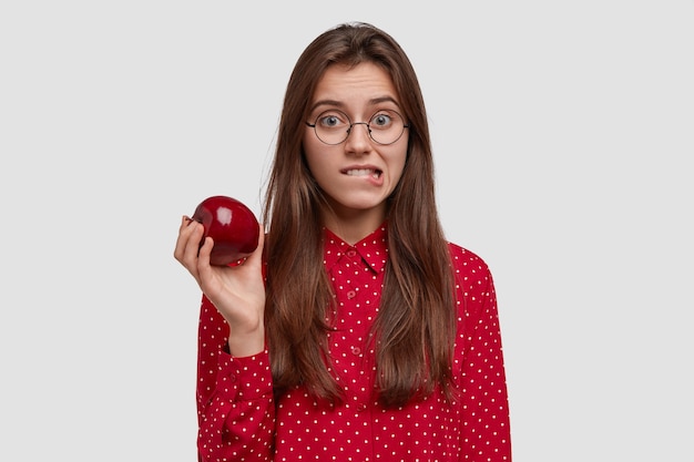 Atractiva mujer joven muerde los labios, los labios inferiores, sostiene la manzana roja fresca, tiene una nutrición saludable, viste una camisa de lunares rojos, se levanta contra el fondo blanco