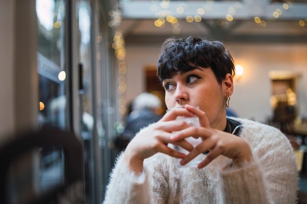 Atractiva mujer joven con pelo corto bebiendo agua en un café y mirando por la ventana