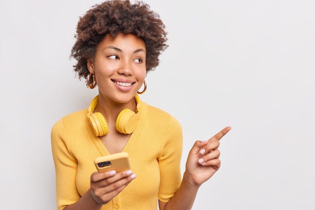 Atractiva mujer joven de pelo rizado con suave sonrisa sostiene modernos auriculares de teléfono móvil alrededor del cuello indica lejos en el espacio de la copia vestida con un jersey amarillo casual aislado sobre una pared blanca