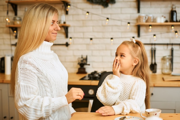 Atractiva mujer joven posando en el elegante y acogedor interior de la cocina decorada con guirnaldas que lleva a su linda niña que está expresando asombro, emocionada con noticias positivas inesperadas, cubriendo la boca