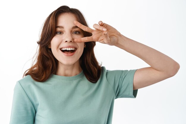 Atractiva mujer joven con rostro perfecto y sonrisa mostrando vsign cerca del ojo y mirando feliz a la cámara manteniéndose positiva de pie contra el fondo blanco