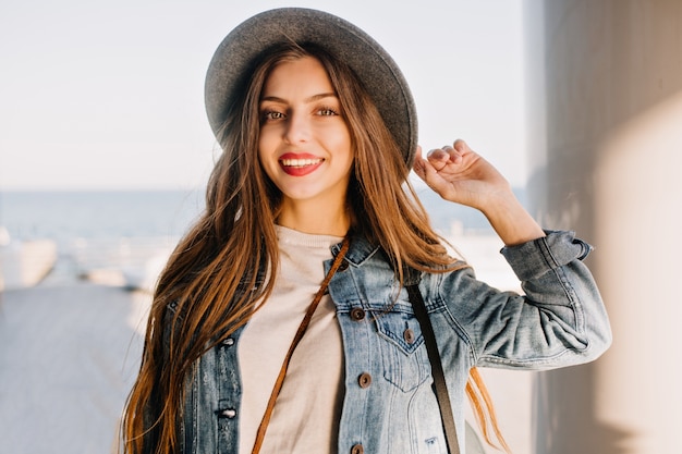 Atractiva mujer joven sensual con sombrero de moda sonriendo y posando con la mano en el fondo borroso.