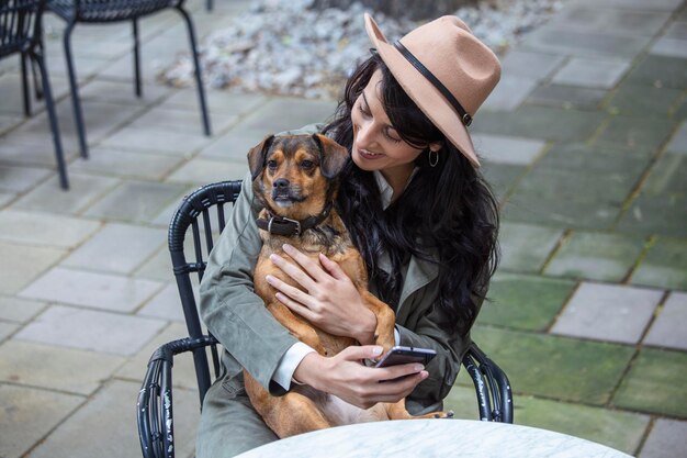 Atractiva mujer joven sentada en una cafetería relajándose y sosteniendo a su perro Cafetería amigable para mascotas hermosa niña con su perro sentado en una cafetería y tomando café