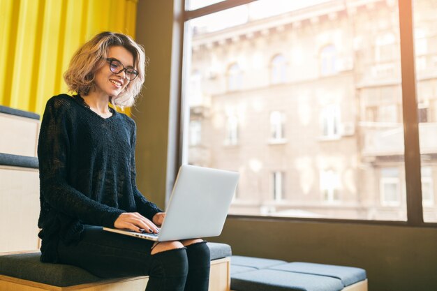 Atractiva mujer joven sentada en la sala de conferencias por ventana grande, trabajando en equipo portátil, con gafas