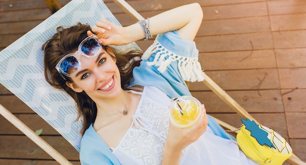 Atractiva mujer joven sentada en la tumbona en traje de moda de verano, estilo hipster, vestido blanco, capa azul, gafas de sol, sonriendo, bebiendo limonada, accesorios elegantes, relajándose en vacaciones