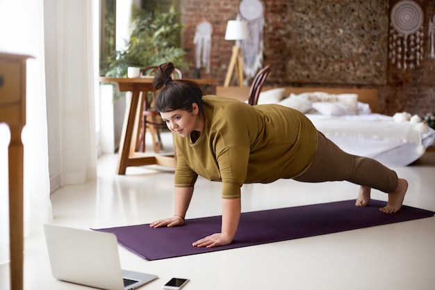 Atractiva mujer joven con sobrepeso descalza haciendo tabla en la estera de yoga mientras entrena en el interior, viendo videos en línea a través de la computadora portátil. Concepto de deporte, bienestar, tecnología y estilo de vida activo y saludable