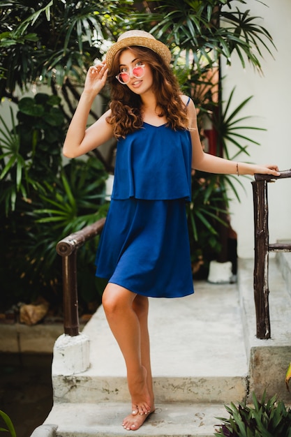 Atractiva mujer joven en vestido azul y sombrero de paja con gafas de sol rosa