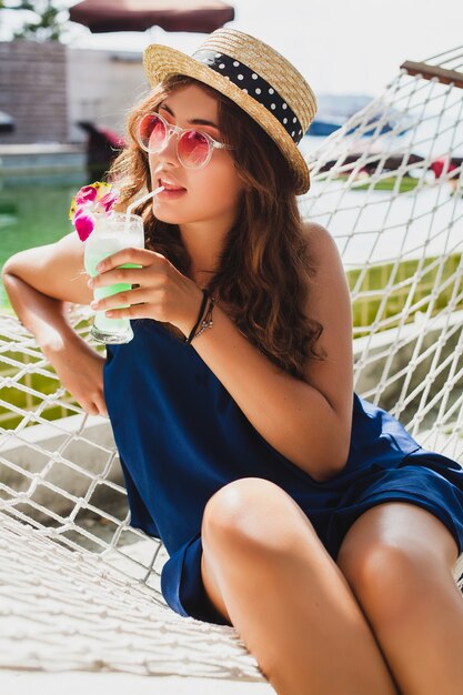 Atractiva mujer joven en vestido azul y sombrero de paja con gafas de sol rosas, bebiendo cócteles de alcohol en vacaciones y sentado en una hamaca