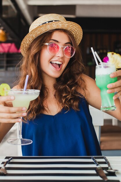 Atractiva mujer joven con vestido azul y sombrero de paja con gafas de sol rosas bebiendo cócteles de alcohol en vacaciones tropicales sentado a la mesa en el bar con traje de estilo veraniego, sonriendo feliz en el estado de ánimo de fiesta