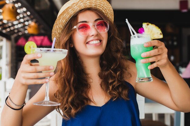 Atractiva mujer joven con vestido azul y sombrero de paja con gafas de sol rosas bebiendo cócteles de alcohol en vacaciones tropicales sentado a la mesa en el bar con traje de estilo veraniego, sonriendo feliz en el estado de ánimo de fiesta