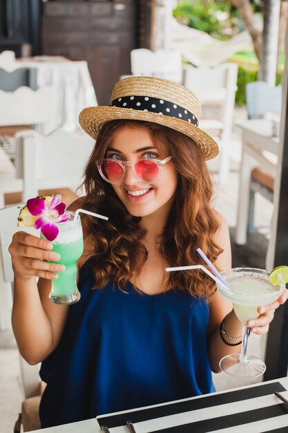 Atractiva mujer joven en vestido azul y sombrero de paja con gafas de sol rosas, bebiendo cócteles de alcohol en vacaciones tropicales y sentado a la mesa en el bar