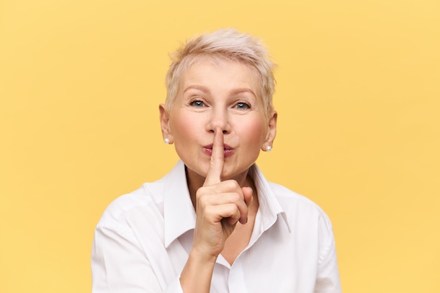 Atractiva mujer de mediana edad con camisa blanca sosteniendo el dedo índice en su boca, haciendo un signo de silencio, diciendo Shh, no le digas a nadie, pidiéndote que guardes su secreto, con una expresión facial misteriosa