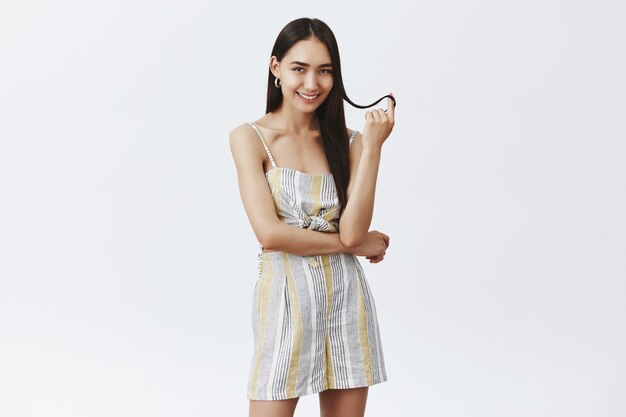 Atractiva mujer de moda y femenina en traje de verano jugando con un mechón de cabello, girándolo con el dedo y sonriendo sensualmente, seduciendo sobre la pared gris