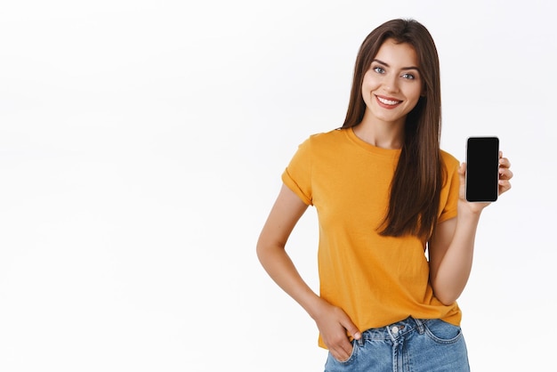 Atractiva mujer moderna y elegante con camiseta amarilla promueve una aplicación móvil con un teléfono inteligente que muestra la pantalla, da un enlace a sus redes sociales de pie, fondo blanco, feliz sonriendo
