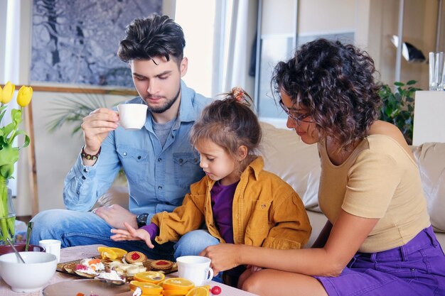 Atractiva mujer morena con anteojos, hombre barbudo con estilo y su linda hijita se sienta en un sofá y almuerza en una sala de estar.