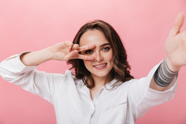 Atractiva mujer morena con camisa blanca toma selfie y muestra el signo de la paz. Niña feliz sonríe ampliamente sobre fondo rosa aislado.